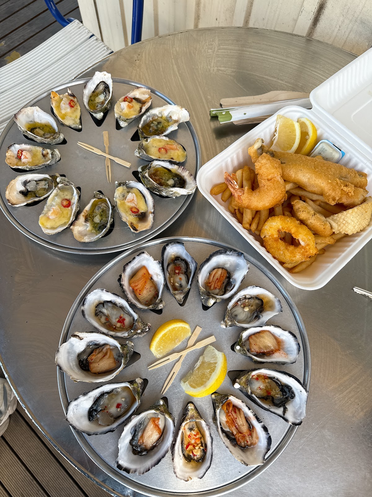 Two platters of dressed oysters beside a box of fish and chips on a steel table